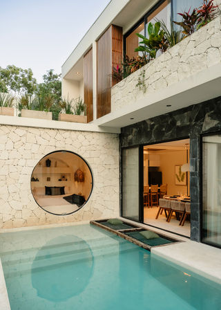 Modern tropical villa courtyard with a turquoise private plunge pool, textured stone walls, circular window into a cozy bedroom, sliding glass doors to a dining area, upper balcony planters and wood accents.