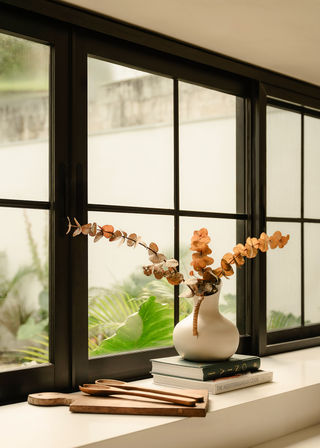 Cozy windowsill vignette with a white ceramic vase of dried eucalyptus on stacked books, wooden cutting boards and spoons, and a black-framed window opening onto a leafy garden.