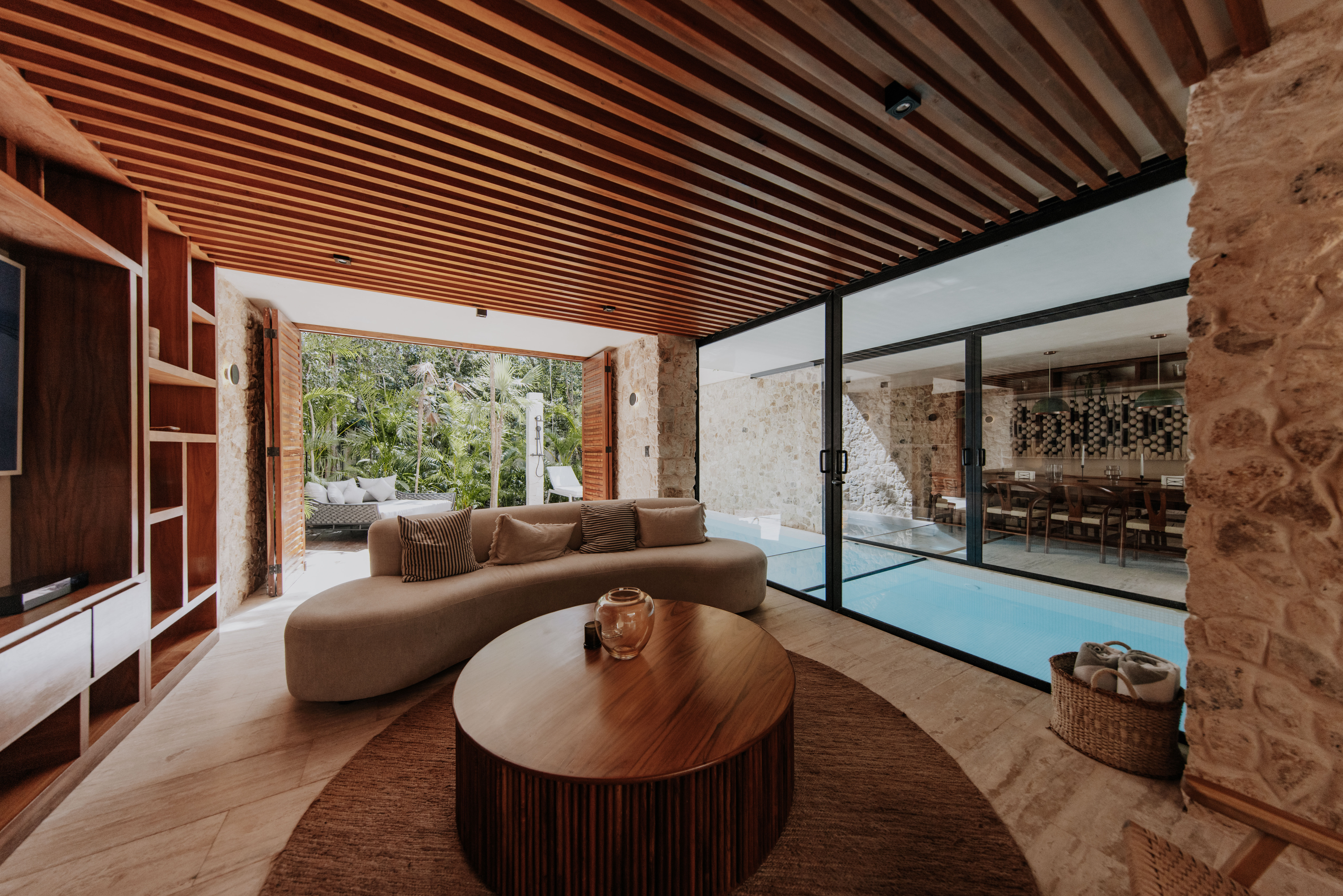 Modern tropical villa lounge with a curved sofa and round wooden coffee table under a wood-slat ceiling, floor-to-ceiling glass doors opening to an indoor pool and dining area, and a lush garden view through open shutters.