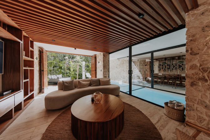 Modern tropical villa lounge with a curved sofa and round wooden coffee table under a wood-slat ceiling, floor-to-ceiling glass doors opening to an indoor pool and dining area, and a lush garden view through open shutters.