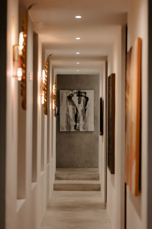 Moody minimalist interior hallway with warm wall sconces and recessed ceiling lights leading to concrete steps and a black-and-white abstract painting at the end.