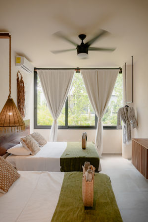 Airy tropical hotel bedroom with two twin beds, green runners, woven pendant light, ceiling fan, tied-back curtains framing lush palms outside and a hanging robe — cozy coastal retreat
