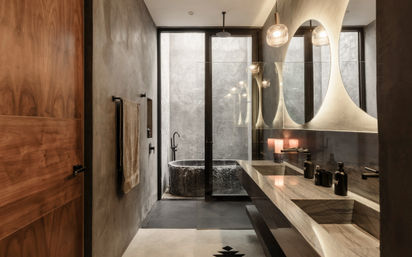 Spa-like modern bathroom with concrete walls, marble double vanity and oval mirrors, glass partition revealing a stone freestanding tub, matte-black fixtures and warm pendant lighting.