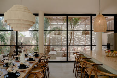 Sunlit modern indoor-outdoor dining space with large black-framed glass wall opening to a shaded tropical patio, wooden dining table set with black plates and glassware, woven pendant lights, bar stools at a kitchen island, and neutral boho decor.