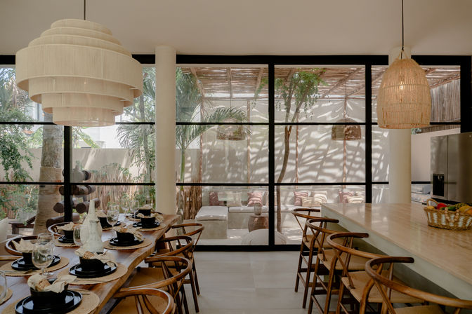 Sunlit modern indoor-outdoor dining space with large black-framed glass wall opening to a shaded tropical patio, wooden dining table set with black plates and glassware, woven pendant lights, bar stools at a kitchen island, and neutral boho decor.