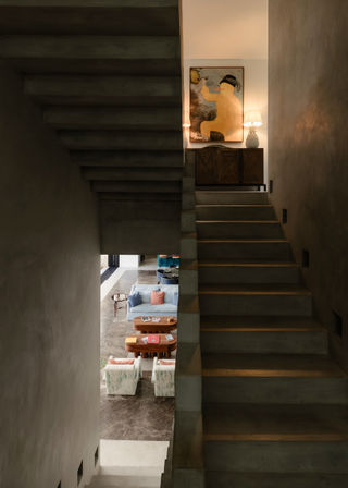 Modern home interior with a sleek concrete staircase framing an open-plan living room below — pastel sofas, wooden coffee tables, marble floor, and a lit console with artwork on the top landing.