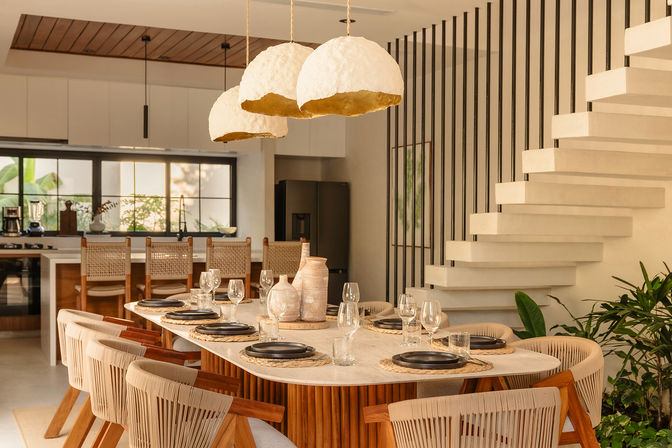 Sunlit modern dining room with a long wooden table set for eight, woven chairs, textured pendant lamps, open kitchen, floating staircase and indoor plants — boho-coastal style.