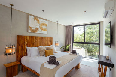 Bright modern bedroom with wooden headboard, neutral linens and pendant lights, large window with leafy tree view, potted plant and neatly folded towels — stylish hotel-style guest room.