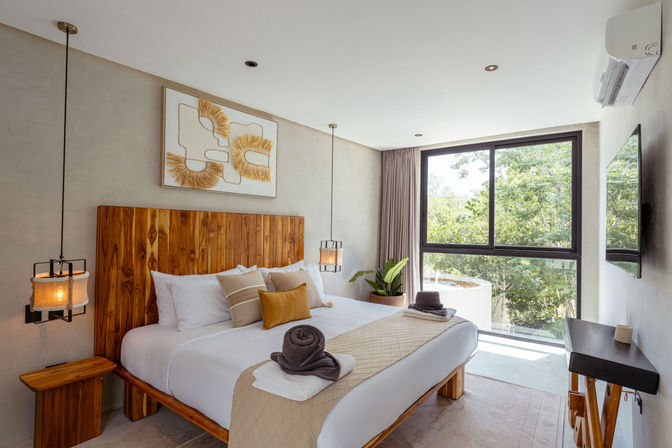 Bright modern bedroom with wooden headboard, neutral linens and pendant lights, large window with leafy tree view, potted plant and neatly folded towels — stylish hotel-style guest room.
