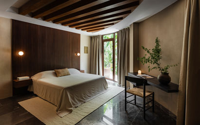 Modern minimalist bedroom with wooden slatted accent wall, king bed in neutral linens, floating nightstands and warm wall sconces, dark marble floor, floor-to-ceiling glass door opening to lush tropical greenery, and a wall-mounted desk with a potted plant.