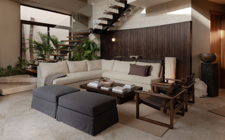 Modern neutral-toned living room interior with cream sectional sofa, dark wood coffee table, leather armchairs, indoor plants, natural stone floor and glass floating staircase.