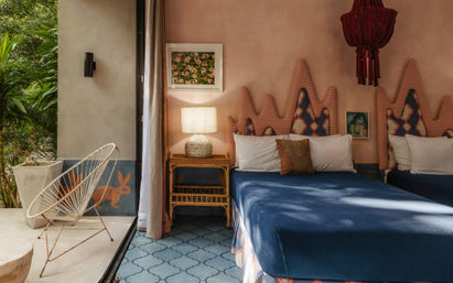 Tropical boutique hotel bedroom with a deep-blue bedspread, scalloped patterned headboards, wicker nightstand and lamp, red hanging lantern, patterned blue tile floor, and an open sliding door to a leafy patio with a white wire lounge chair.