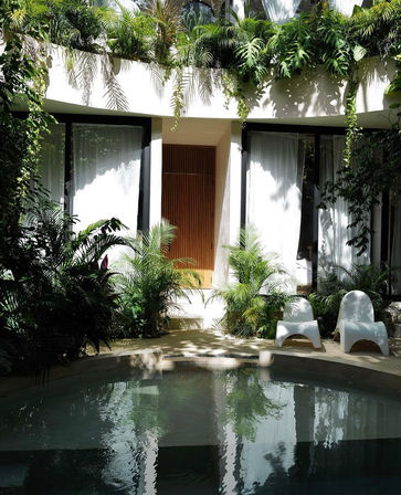 Tropical courtyard with a circular plunge pool reflecting a modern white villa facade, wooden slatted door, lush hanging and potted palms, and two white sculptural lounge chairs.