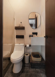 Cozy compact modern bathroom with white one-piece toilet, wall-mounted plaster sink, organic oval mirror, dark wood floating shelves, woven storage basket and charcoal square floor tiles.