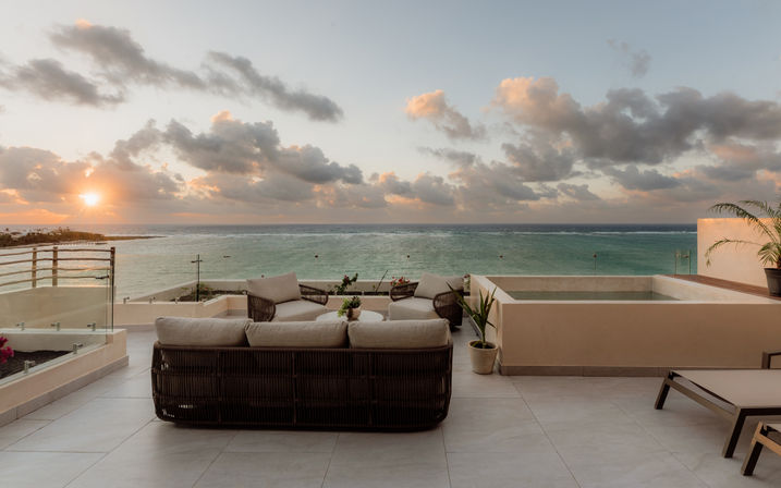 Oceanfront rooftop terrace at sunset with wicker lounge sofa and chairs, potted plants and a small plunge pool overlooking turquoise sea and a dramatic cloud-filled sky.