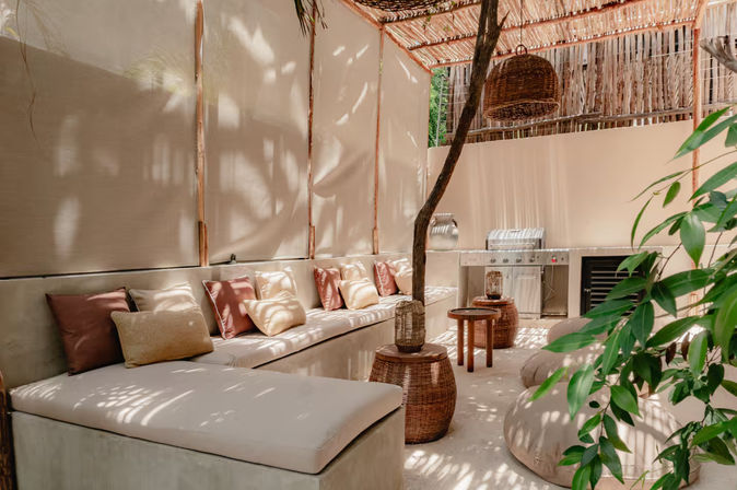 Sun-dappled boho outdoor lounge patio with a long built-in cushioned bench and blush-beige pillows, wicker pendant light and side tables, round floor poufs, stainless-steel grill and potted greenery under a bamboo pergola.