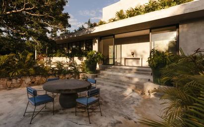 Sunlit tropical outdoor dining terrace at a modern concrete villa with a round stone table, four blue-cushioned metal chairs, stone steps up to glass doors, and lush potted greenery.