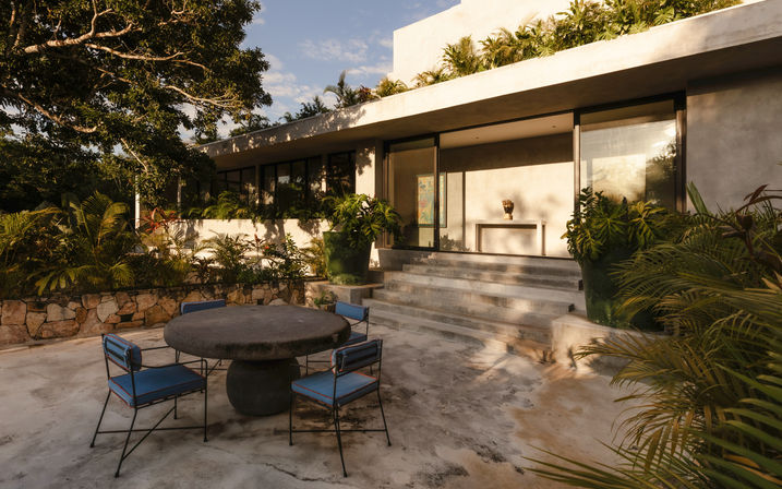 Sunlit tropical outdoor dining terrace at a modern concrete villa with a round stone table, four blue-cushioned metal chairs, stone steps up to glass doors, and lush potted greenery.
