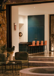 Cozy modern patio opening to an illuminated indoor bar with deep teal cabinets, four coral bar stools, stone accent wall, potted plants, low outdoor lounge chairs and a circular fire pit under warm evening lighting