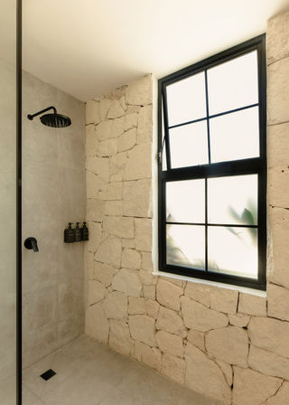 Sunlit rustic stone-walled shower with matte-black rain showerhead, wall-mounted black dispensers and a frosted grid window