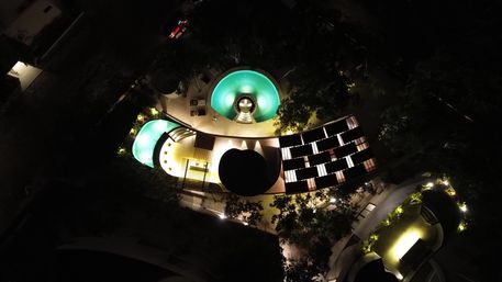 Aerial night view of glowing turquoise circular resort pools and curved terraces with illuminated pathways and lush landscaping in a hotel courtyard.