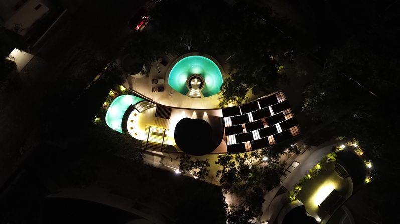 Aerial night view of glowing turquoise circular resort pools and curved terraces with illuminated pathways and lush landscaping in a hotel courtyard.
