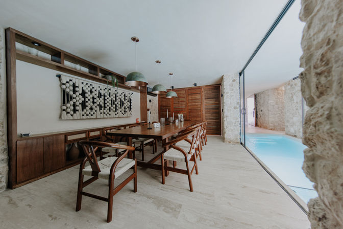 Modern rustic dining room with a long wooden table and woven-seat chairs under green pendant lights, stone walls and sliding glass doors opening to a bright indoor pool