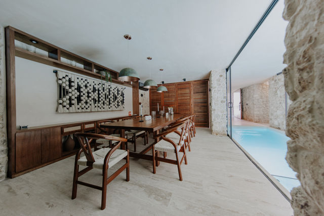 Modern rustic dining room with a long wooden table and woven-seat chairs under green pendant lights, stone walls and sliding glass doors opening to a bright indoor pool