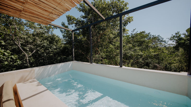 Sunlit private terrace plunge pool with pale blue water, bamboo pergola and glass railing overlooking a lush tropical jungle canopy