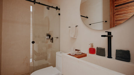 Sleek neutral-toned modern bathroom with glass sliding shower door, round wall mirror, matte-black tall faucet, white vanity with wooden tray holding a glass and red soap dispenser, toilet, and neatly folded gray towels.
