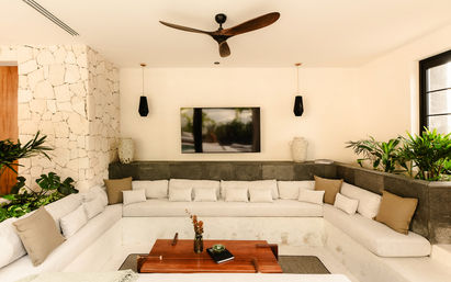 Airy modern tropical living room with U-shaped cream built-in sofa, stone accent wall, wooden ceiling fan, wall-mounted TV, wooden coffee table and lush indoor plants.