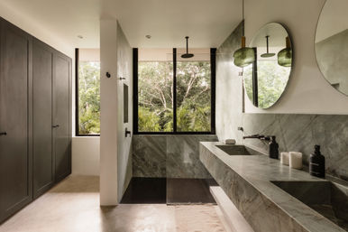 Sunlit modern marble bathroom with double sinks, round mirrors, walk-in shower and large window overlooking lush tropical greenery