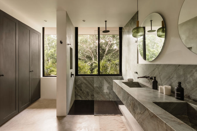 Sunlit modern marble bathroom with double sinks, round mirrors, walk-in shower and large window overlooking lush tropical greenery