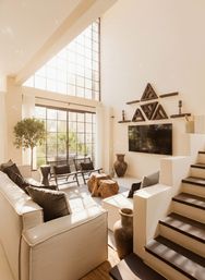 Sunlit modern loft living room with floor-to-ceiling grid windows, neutral sectional sofa, wooden stump coffee tables, indoor tree, staircase, wall-mounted TV and geometric wood wall art.