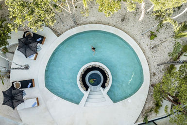 Aerial view of a circular turquoise resort pool with a built-in round seating nook and white steps, one swimmer in the water, black woven parasols and sun loungers on a sandy tropical patio surrounded by palm trees — perfect tropical getaway scene.