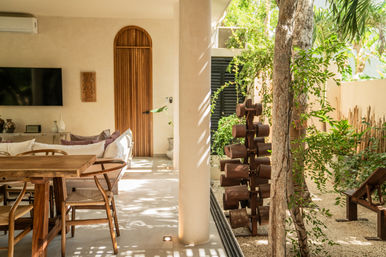 Sunlit tropical villa indoor-outdoor living area with wooden dining table and chairs, comfy sofa, arched wood door, column, and a lush garden patio with stacked wooden sculpture and gravel.