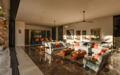 Sunlit open-plan living room in a modern tropical villa with polished marble floors, low gray sofas and coral pillows, wooden coffee tables, colorful armchair, dining area with orange chairs, and large folding glass doors opening to a stone patio.