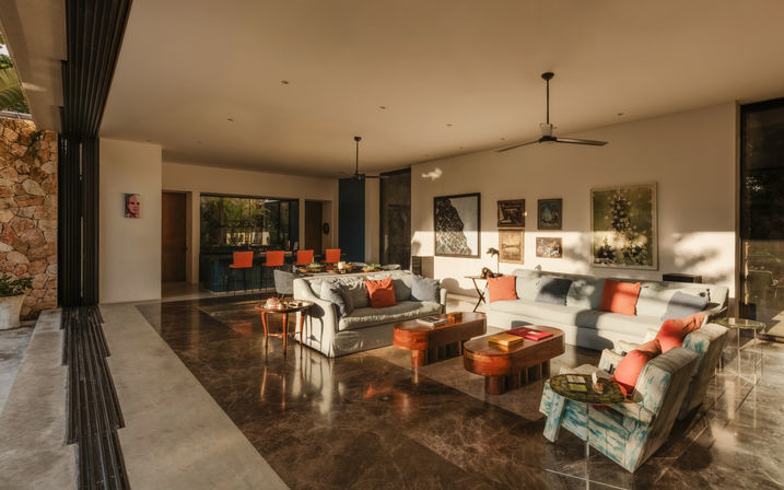 Sunlit open-plan living room in a modern tropical villa with polished marble floors, low gray sofas and coral pillows, wooden coffee tables, colorful armchair, dining area with orange chairs, and large folding glass doors opening to a stone patio.