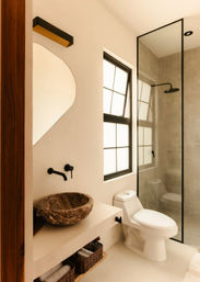 Sunlit modern bathroom with round stone vessel sink, matte black wall-mounted faucet, organic-shaped mirror, one-piece white toilet and glass shower with black frame, woven storage baskets and warm neutral finishes.