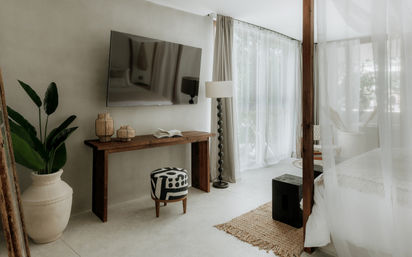 Airy sunlit bedroom interior with wall-mounted TV above a rustic wooden console, patterned ottoman, tall potted tropical plant, floor lamp, sheer floor-to-ceiling curtains and woven rug — relaxed coastal-inspired decor.