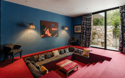 Cozy sunken lounge with red carpet and built-in gray seating, blue painted walls, patterned throw pillows, woven ottoman, wall sconces and abstract art, plus floor-to-ceiling glass doors opening to a stone courtyard