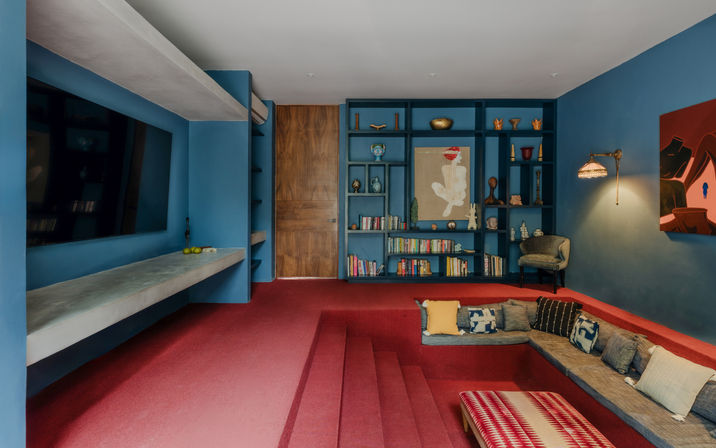 Sunken living room with deep red carpet and blue walls, built-in shelving filled with books and decor, wooden door, large wall-mounted TV, stepped seating nook with assorted throw pillows, patterned ottoman and warm wall sconce lighting.