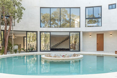 Modern minimalist villa courtyard with a circular turquoise pool and sunken round island lounge, floor-to-ceiling glass walls reflecting surrounding trees and a floating interior staircase