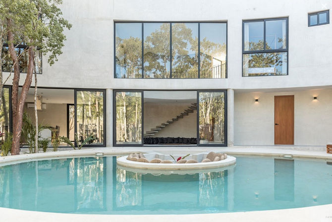 Modern minimalist villa courtyard with a circular turquoise pool and sunken round island lounge, floor-to-ceiling glass walls reflecting surrounding trees and a floating interior staircase