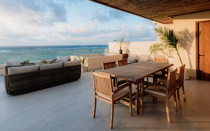 Sunlit oceanfront terrace with wooden dining table and chairs, cushioned wicker sofa, potted palms and glass railing overlooking a turquoise sea at golden hour.