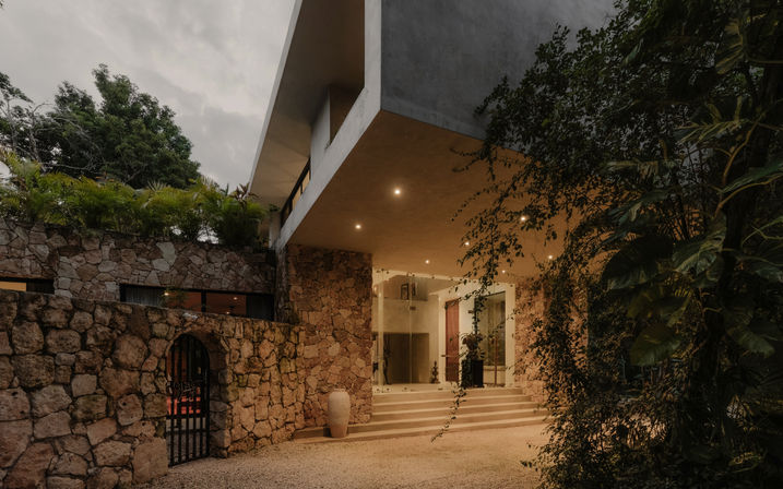 Modern stone-clad house entrance with cantilevered concrete upper level, warm recessed lighting on steps, a decorative vase and lush tropical greenery at dusk.