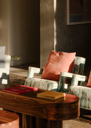 Cozy sunlit living room featuring a distressed teal patterned armchair with a coral cushion, a polished wooden coffee table with red and yellow books, and soft neutral shadows.