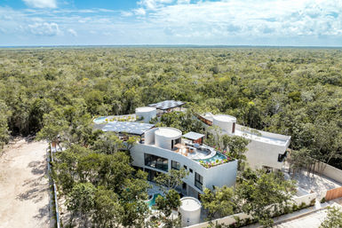Aerial view of a modern eco-friendly villa with rooftop pool and solar panels nestled in a dense tropical forest, ocean visible on the distant horizon.
