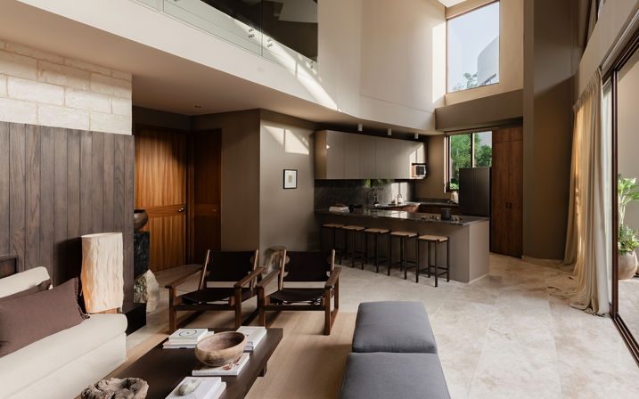 Sunlit modern open-plan living room and kitchen with double-height ceiling, stone tile floor, warm wood accents, breakfast bar with stools, lounge chairs, and sliding glass doors to greenery.