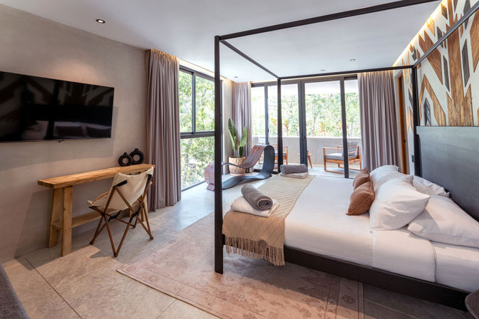Sunlit modern resort-style bedroom with four-poster canopy bed, neutral linens and rolled towels, wall-mounted TV, wooden desk and sculptural lounge chair, floor-to-ceiling glass doors leading to a balcony with lush green views and outdoor seating.